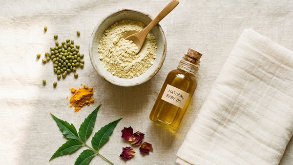 Flatlay of natural baby skin care essentials — herbal bath powder, cold-pressed oil, turmeric and moong, and a soft towel on cream linen